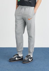 Pelēki treniņbikses ar oranžu Nike logo, ar auklu jostas vietā un elastīgām piedurknēm, apvienotas ar melniem Nike sporta apaviem un baltām potīšu zeķēm.