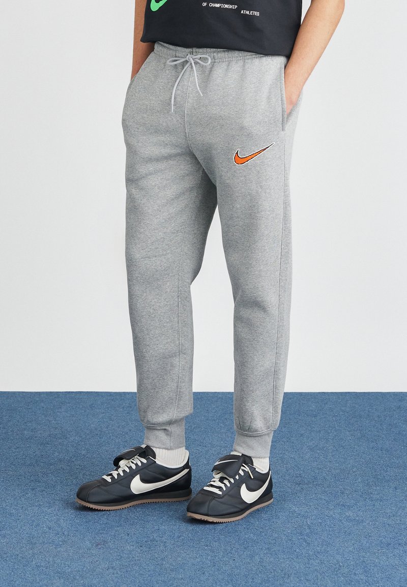Pelēki treniņbikses ar oranžu Nike logo, ar auklu jostas vietā un elastīgām piedurknēm, apvienotas ar melniem Nike sporta apaviem un baltām potīšu zeķēm.