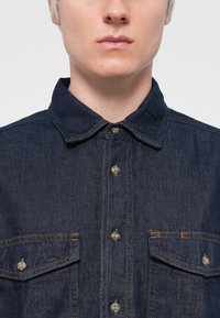 Mørk denimskjorte med spredd krage, to frontlommer og kontrastsømming. Knappefront med plastknapper.