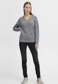 Grauer Strickpullover mit V-Ausschnitt und gerippten Bündchen, kombiniert mit schwarzen engen Jeans und grauen Sneakers, vor einem einfarbigen Hintergrund.