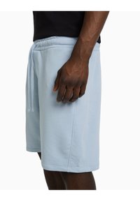 Muž oblečený v světle modrých šortkách na stažení a černém tričku, stojící s rukou uvolněně vedle těla na bílém pozadí.