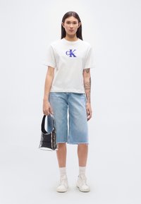 Λευκό βαμβακερό T-shirt με μπλε λογότυπο Calvin Klein, συνδυασμένο με μπλε denim culottes και λευκά αθλητικά παπούτσια. Κρατά μια μαύρη λαμπερή τσάντα χειρός.