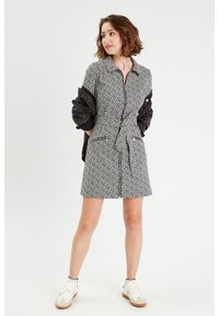 Robe à manches courtes, à motifs noir et blanc, avec une fermeture éclair à l'avant, une ceinture à la taille et deux poches latérales. Portée avec une veste sombre et des baskets.