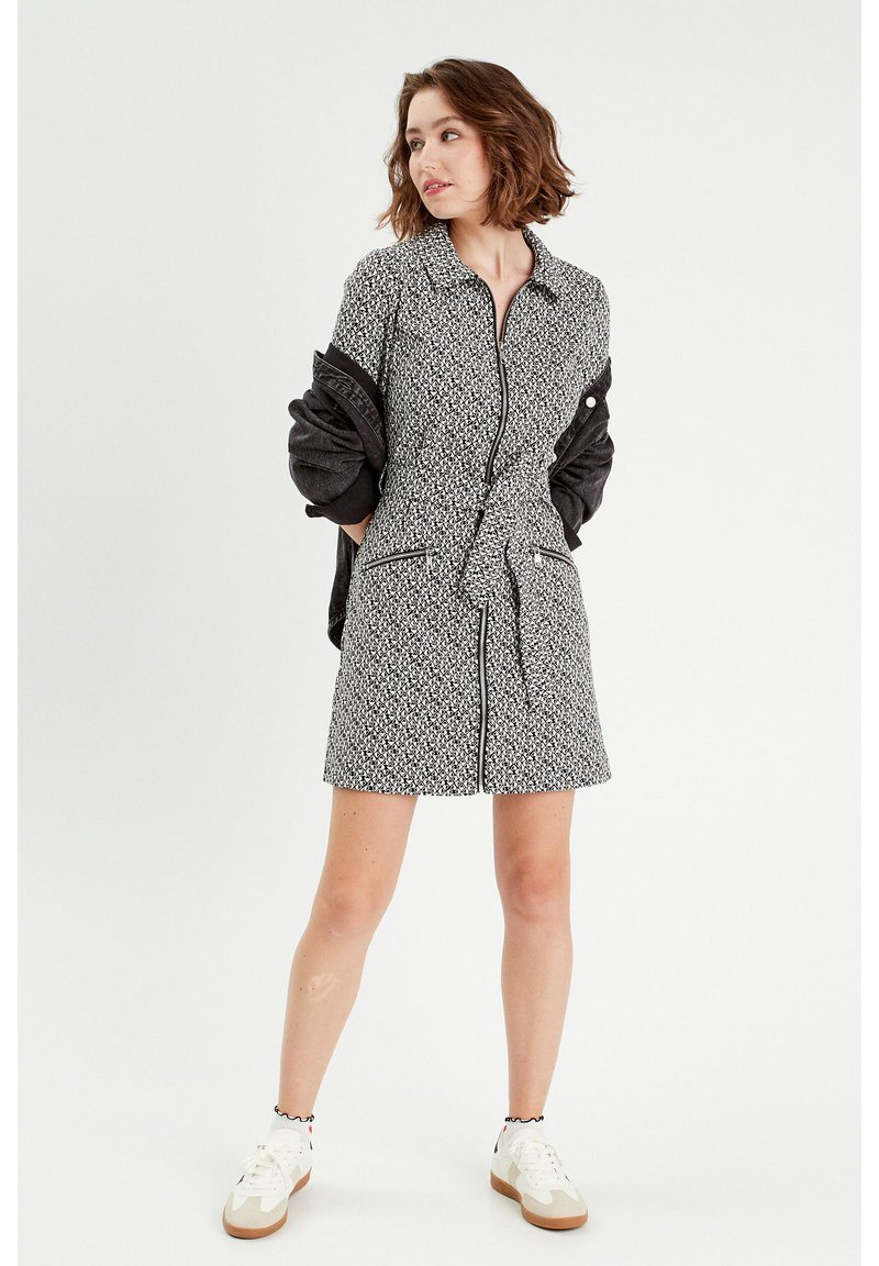 Robe à manches courtes, à motifs noir et blanc, avec une fermeture éclair à l'avant, une ceinture à la taille et deux poches latérales. Portée avec une veste sombre et des baskets.