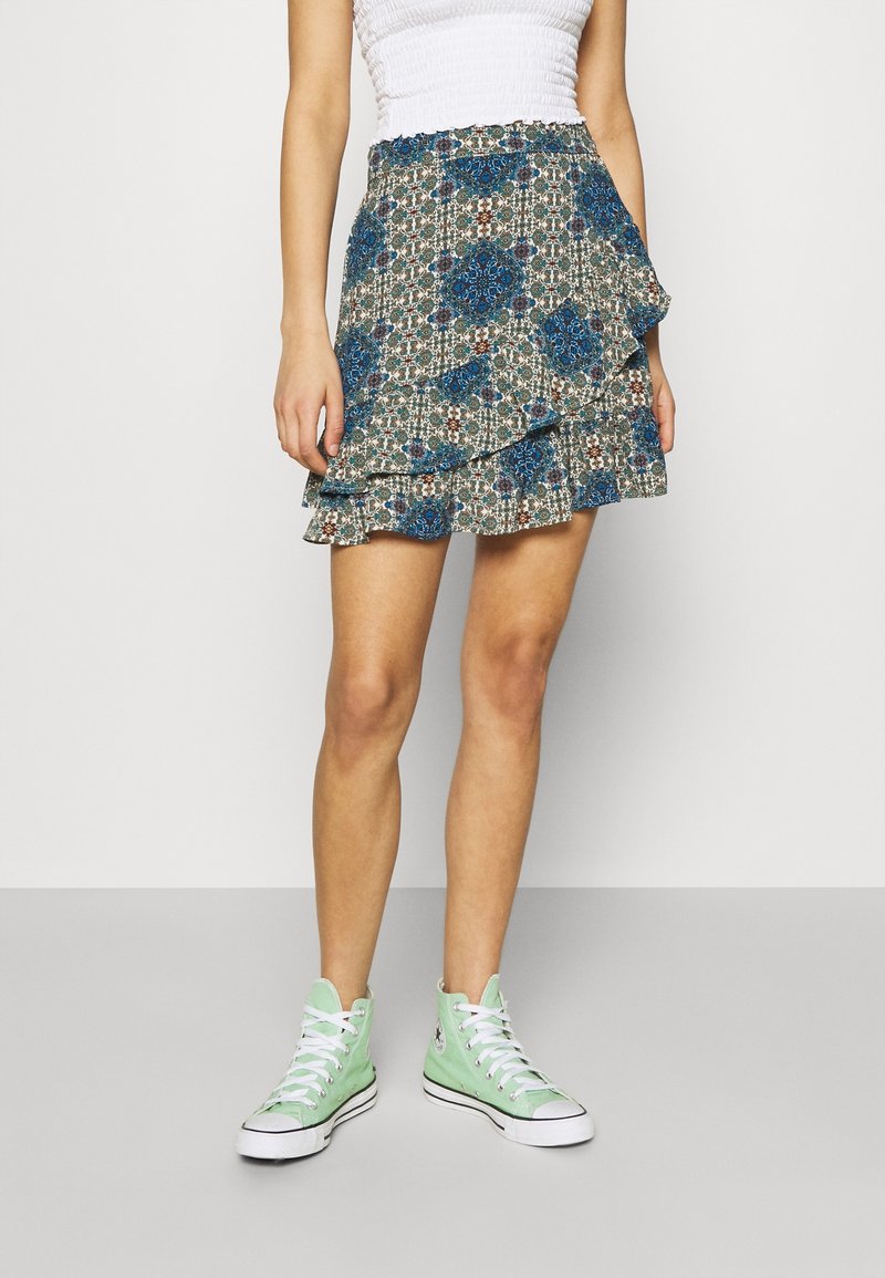 Femme portant une mini-jupe à volants à motifs bleus et verts, un haut blanc froncé et des baskets montantes vert clair, debout sur un sol gris.