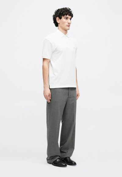 Biele polo tričko s krátkymi rukávmi a jemným logom. Kombinované s ľahkými sivými, na mieru šitými nohavicami a čiernymi spoločenskými topánkami.