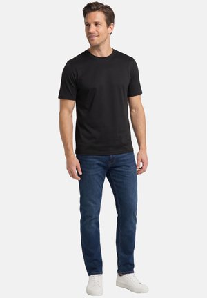 Ein Mann steht, trägt ein schwarzes T-Shirt mit kurzen Ärmeln, blaue Jeans und weiße Turnschuhe, blickt leicht nach links mit neutralem Hintergrund.