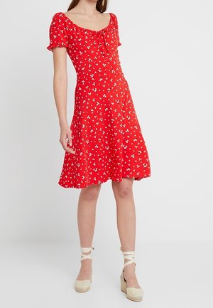 Kobieta ubrana w czerwone, krótkie rękawy, sukienkę do kolan z małym białym kwiatowym wzorem oraz beżowe koturny espadryle z wiązaniami wokół kostek.