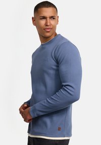 Blauw geribd gebreid shirt met lange mouwen, een ronde halslijn en aangesloten manchetten. Heeft een klein logo detail en een contrasterende witte zoom aan de taille.