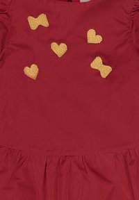 Indumento rosso con orlo inferiore arricciato, caratterizzato da cuori e fiocchi gialli ricamati di varie dimensioni sulla parte anteriore. Texture liscia.