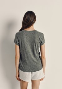 Moteris su ilgais rudais plaukais stovi veidu į smėlio spalvos sieną, vilkėdama pilką trumpomis rankovėmis palaidinę ir šviesios spalvos šortus.