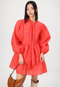 Korálová šaty s puff-sleeve rukávmi z textúrovanej látky, s detailom na viazanie v oblasti krku a rozšírenou sukňou. V kombinácii s hnedou kabelkou s embosovaným hadím vzorom a bielymi čižmami.