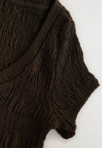 Tissu texturé brun foncé avec un motif froissé, comportant une manche courte et un col rond sur un fond clair.