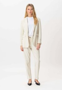 Blond kvinde iført en crèmeguldragt med en hvid skjorte og sorte spidse stiletter, der står mod en hvid baggrund.