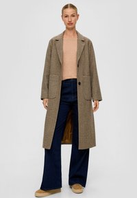 Vrouw staat in een lange bruine geblokte jas, beige trui, donkere wijde spijkerbroek en tan platform schoenen tegen een effen achtergrond.