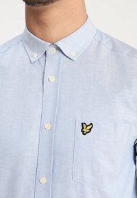 Camisa azul claro abotonada, hecha de tela suave, con un bolsillo en el pecho que tiene un logo bordado en amarillo y negro.