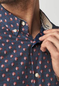Close-up van een persoon die de kraag van een blauw shirt met een rood-wit bloemenpatroon en witte knopen aanpast.