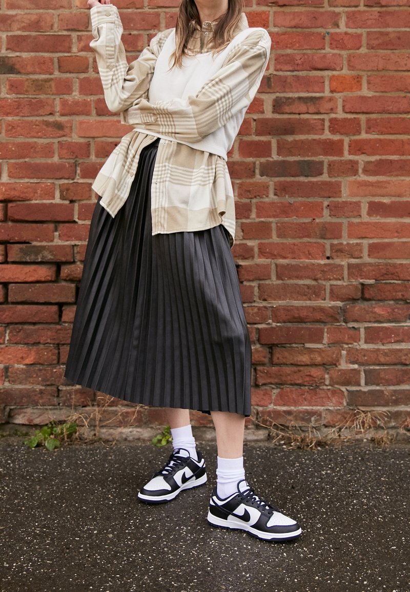 Chemise à manches longues à carreaux beige superposée sur un haut blanc, associée à une jupe midi plissée noire et des baskets noir et blanc avec des chaussettes blanches.