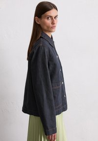 Dark denim jacket with a button front, pointed collar, and two side pockets. Features contrast stitching and a relaxed fit, paired with a pleated skirt.