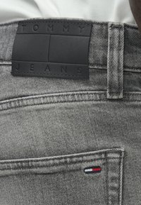 Vaqueros de mezclilla gris con un parche de cuero negro etiquetado "TOMMY JEANS" y un pequeño detalle bordado en rojo, blanco y azul.