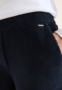 Pantaloni in corduroy blu navy con righe verticali, dotati di una cintura liscia, tasche laterali e un piccolo tag metallico del marchio.