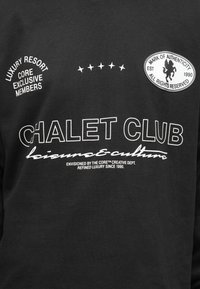 Tessuto nero con testo bianco che legge "CHALET CLUB leisure & culture," icone a stella e distintivi che dicono "Luxury Resort Core Exclusive Members" e "Mark of Authenticity."