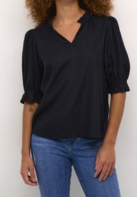Blouse noire à manches courtes avec un décolleté en V et des manches bouffantes froncées. Tissu lisse avec une légère brillance. Portée avec un jean bleu.