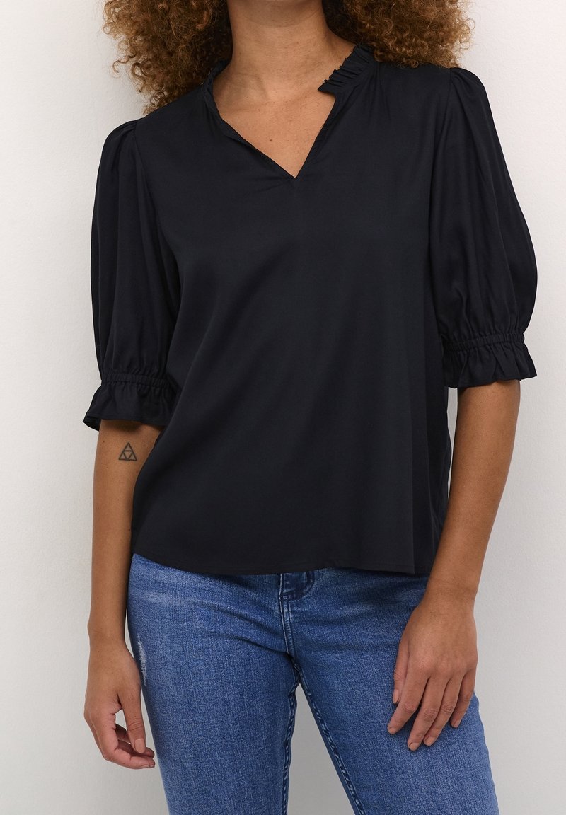 Blouse noire à manches courtes avec un décolleté en V et des manches bouffantes froncées. Tissu lisse avec une légère brillance. Portée avec un jean bleu.