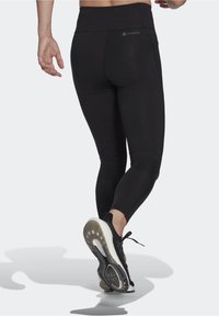 Svarta träningsleggings med hög midja, slät textur och en dold ficka. Modellen bär svarta löparskor med vit sula.