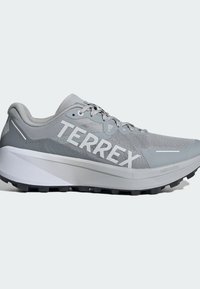 Zapatilla deportiva gris con parte superior de malla, un destacado logotipo "TERREX", una gruesa entresuela blanca y una suela negra texturizada para mayor tracción.