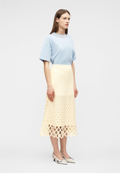 Femme debout portant un haut à manches courtes bleu clair, une jupe midi texturée couleur crème avec des découpes géométriques, et des escarpins blancs à bride arrière.