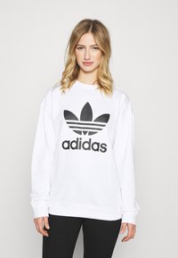 Bijela dukserica s velikim crnim Adidas logotipom i dizajnom s tri pruge. Mekani materijal s dugim rukavima i opuštenim krojem.