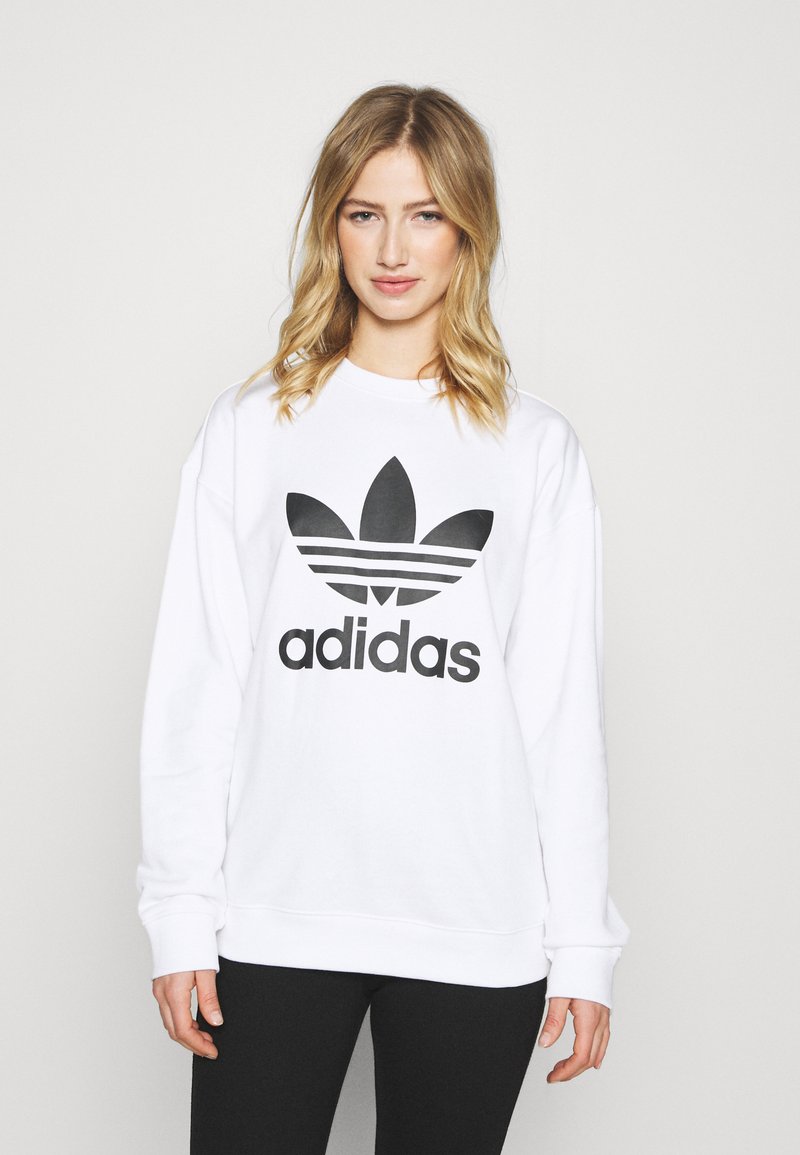 Bijela dukserica s velikim crnim Adidas logotipom i dizajnom s tri pruge. Mekani materijal s dugim rukavima i opuštenim krojem.