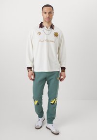 Long-sleeve white polo with burgundy collar and embroidered text; teal sweatpants with yellow accents and graphics; white and brown sneakers.
