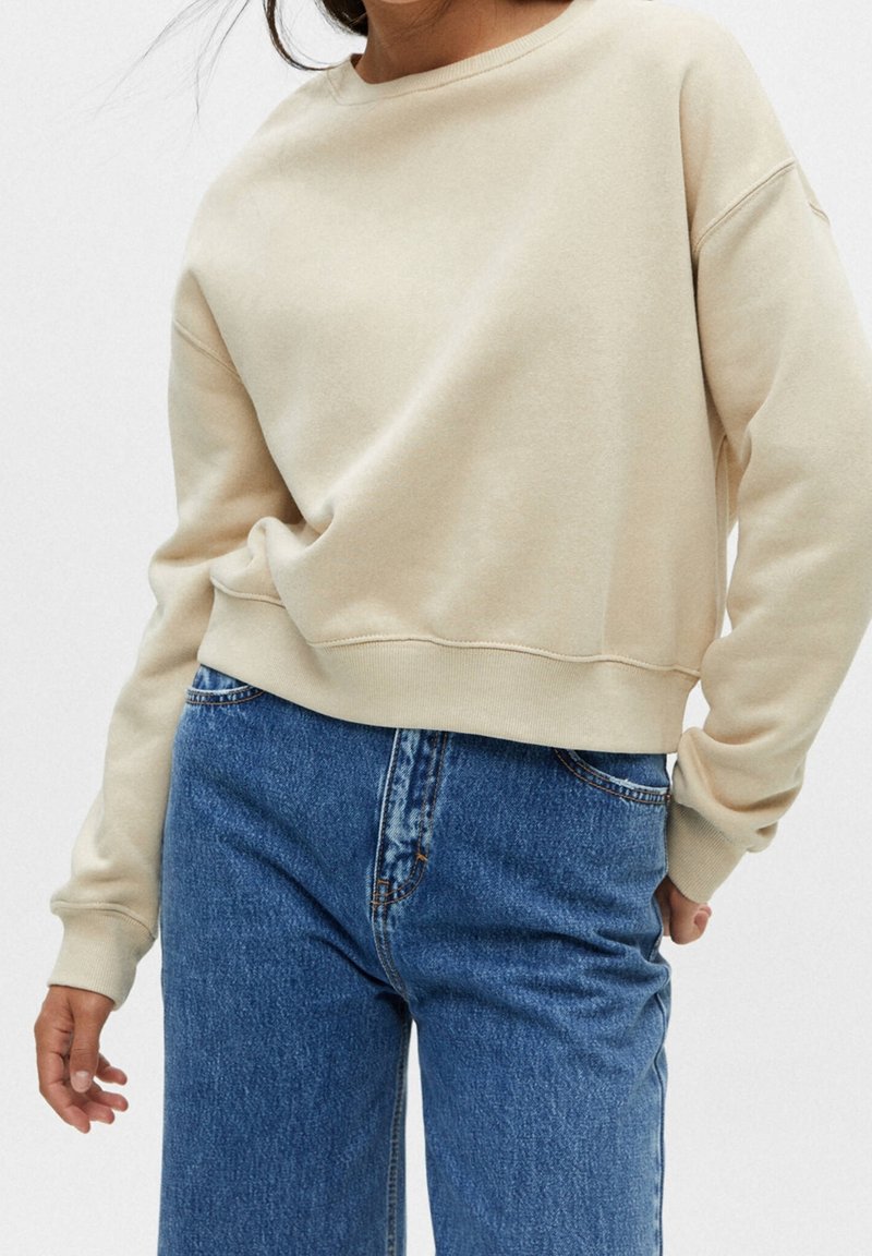 Personne portant un sweat-shirt beige à manches longues et un jean bleu taille haute, debout avec une main dans la poche et l'autre sur le côté.