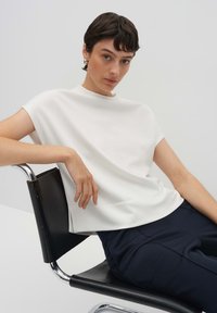 Haut blanc à manches courtes avec un col montant, texture lisse, assorti à un pantalon bleu marine foncé. Modèle assis sur une chaise noire.