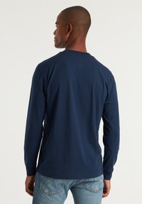 Marineblaue Langarm-T-Shirt mit Rundhalsausschnitt, aus Baumwollstoff und einer flachen Nahtdetails auf der Rückseite. Kombiniert mit hellblauen Jeans.