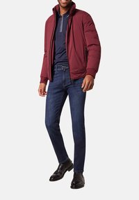 Veste rembourrée bordeaux avec un col montant, associée à un polo bleu marine et un jean en denim foncé. Des bottes de cheville en cuir noir complètent la tenue.