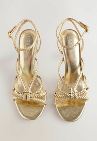 Guld högklackade sandaler med remmar, flätad detaljering och en glänsande yta. Har en platt tånava och justerbara ankelsremmar.