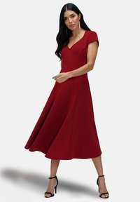 Mid-length red dress with a fitted bodice, cap sleeves, and a flared skirt. Smooth texture and deep V-neckline detail. Black high-heeled sandals.