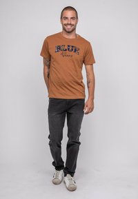 T-shirt marron à manches courtes avec "BLUE & Jeans" imprimé en noir et à carreaux. Porté avec un jean foncé et des baskets blanches. Tatouage visible sur le bras.