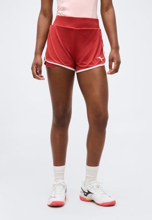 Αθλητής που φοράει κόκκινα αθλητικά σορτς με λευκή επένδυση και λογότυπο, λευκές κάλτσες και λευκά αθλητικά παπούτσια με μαύρες και ροζ λεπτομέρειες.