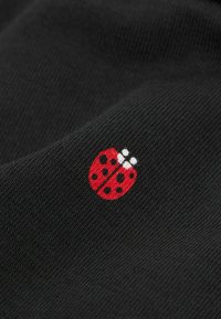 Czarna teksturowana tkanina z małym haftem czerwonej biedronki z czarnymi kropkami i białymi akcentami, umieszczona w dolnym prawym rogu.