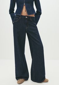 Person wearing dark blue wide-leg jeans and a matching cropped denim jacket, standing with hands in pockets on a plain background.
