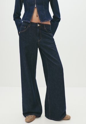 Personne portant un jean large bleu foncé et une veste en denim assortie, debout avec les mains dans les poches sur un fond uni.