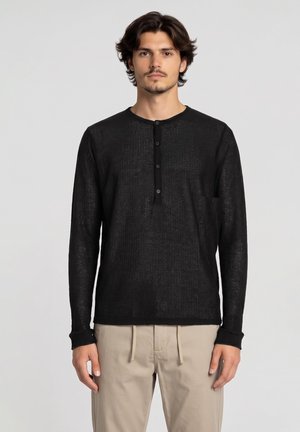 Man met halflang donker haar, gekleed in een zwart langmouwig henley-shirt en beige broek met trekkoord, staand voor een effen achtergrond.