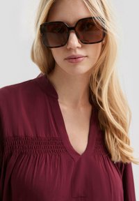 Bordeauxfarbige Bluse mit gerafften Details an der Brust, V-Ausschnitt und subtiler Schimmer. Modell trägt große, dunkel gefärbte Sonnenbrille in quadratischer Form.