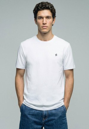 Jeune homme aux cheveux courts et bouclés portant un t-shirt blanc uni avec un petit logo sur la poitrine et un jean bleu, debout les mains dans les poches.