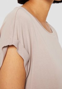 Světle růžový top s krátkým rukávem, kulatým výstřihem a uvolněným střihem. Tkanina vypadá hladce, má jemnou texturu a měkce stočené okraje rukávů.