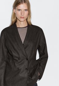 Žena s rovnými blond vlasy, oblečená v tmavém dvouřadém blejzru přes pruhovanou košili, na jednobarevném pozadí.